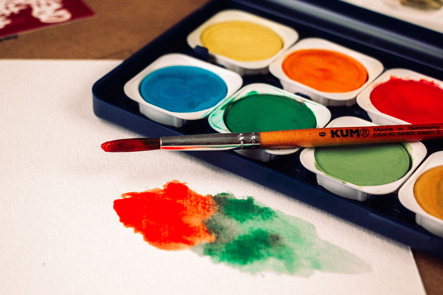 Runder KUM Feinhaar Aquarellpinsel mit roter Farbe über einem Farbmalkasten, daneben ein Aquarellverlauf in Rot und Grün